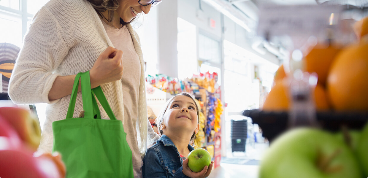 The image depicts a woman holding a bag in her arm and a kid holding a green apple in his hand. They are both watching and sharing a smile.